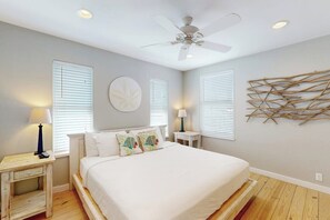 Une chambre à coucher comprenant un lit, deux tables de chevet, un ventilateur de plafond et une œuvre d’art accrochée au mur.