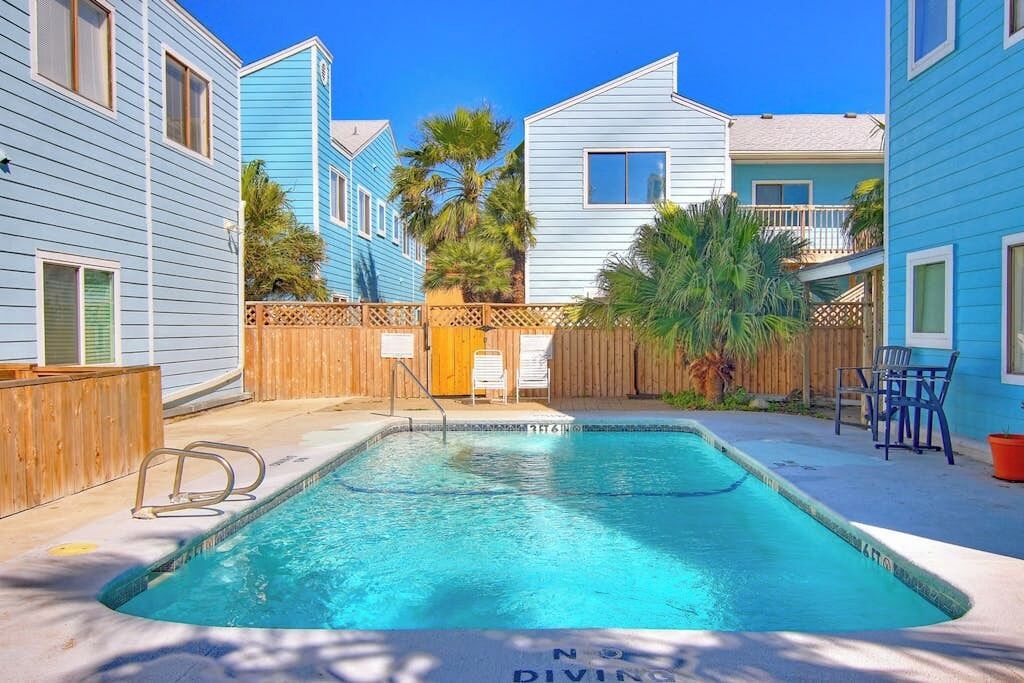 A swimming pool with a wooden deck, surrounded by buildings with blue siding and a wooden fence.