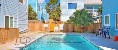 A swimming pool with a wooden deck, surrounded by buildings with blue siding and a wooden fence.