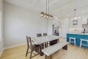 A modern kitchen with a dining area, featuring a large wooden table, bar stools, and pendant lighting. - Roman Empire - Fenced Yard & Patio Near the Bay (Corpus Christi)