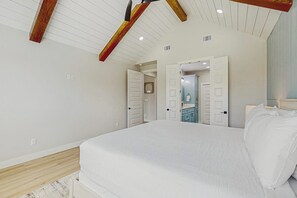 Ein Schlafzimmer mit einem großen Bett, Holzbalken an der Decke und Blick in einen angrenzenden Raum.