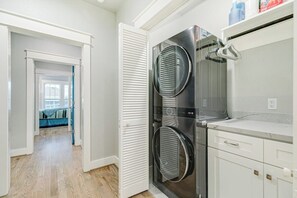 Une buanderie équipée de deux sèche-linge superposés, d’un plan de travail blanc et d’une porte donnant sur une chambre.