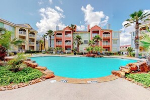 A hotel with a swimming pool surrounded by palm trees and a clear sky. - Dawn Beach Condominiums #323 (Galveston)