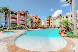 A resort with a large swimming pool surrounded by multi-story buildings with balconies, palm trees, and a clear sky. - Dawn Beach Condominiums #323 (Galveston)