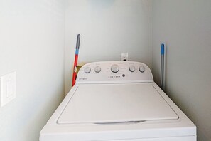 A front-loading Whirlpool washer with a control panel, a red and blue mop, and a white wall socket. - Dawn Beach Condominiums #323 (Galveston)