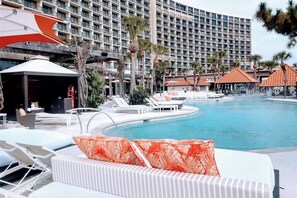 A hotel pool area with lounge chairs, a swimming pool, and a large building in the background. - Sunset Dream - Gulf-Front, Pools & Pickleball (Galveston)