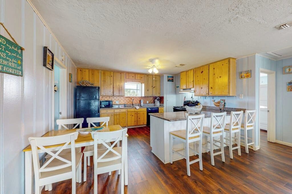 Een keuken met houten kastjes, een centraal eiland met barkrukken, en een eettafel met stoelen.