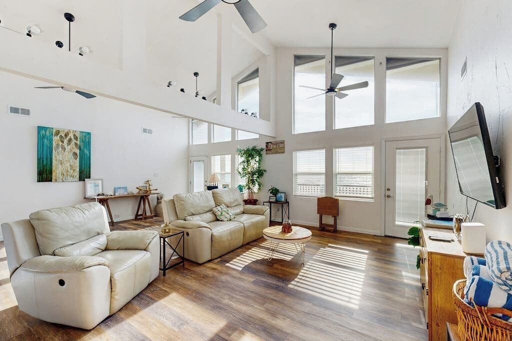 A spacious living room with a high ceiling, large windows, and a ceiling fan.