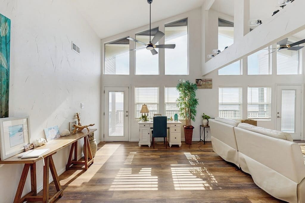 A spacious room with a high ceiling, large windows, a ceiling fan, a desk with a chair, a sofa, and a potted plant.
