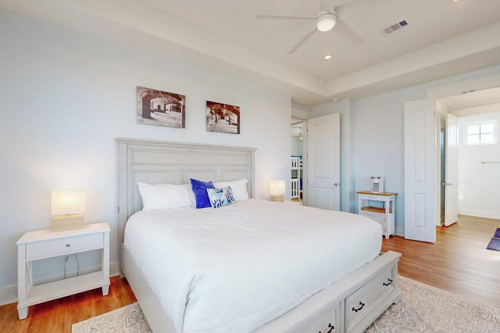 A bedroom with a large bed, white nightstands, a ceiling fan, and artwork on the wall.