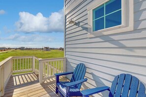 A wooden deck with two Adirondack chairs, a white railing, and a view of a green field. - Sunset Bay - Bay Views, 2 Balconies, Sleeps 12 (Galveston)