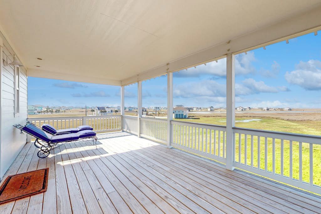 A covered outdoor area with a wooden deck, two lounge chairs, and a view of a grassy field and distant buildings.