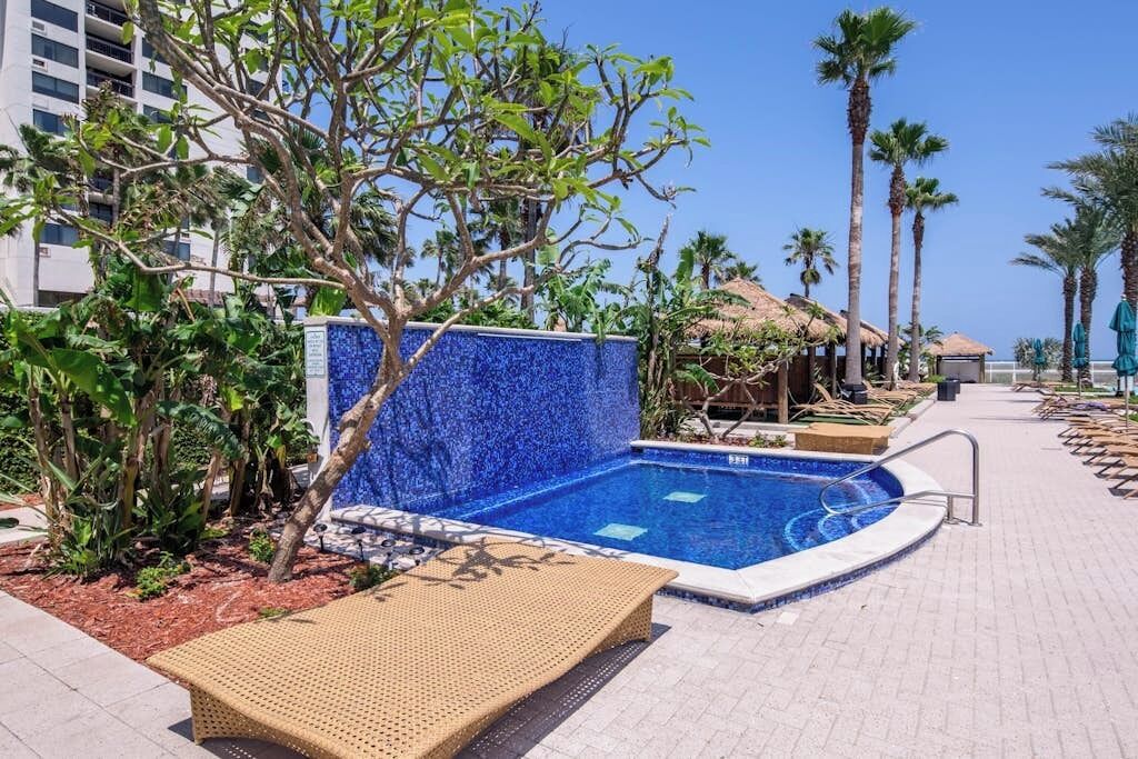 Un área de piscina con una pared de mosaico azul, sillas de descanso, palmeras y un edificio al fondo.