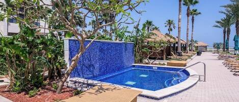 Un área de piscina con una pared de mosaico azul, sillas de descanso, palmeras y un edificio al fondo.