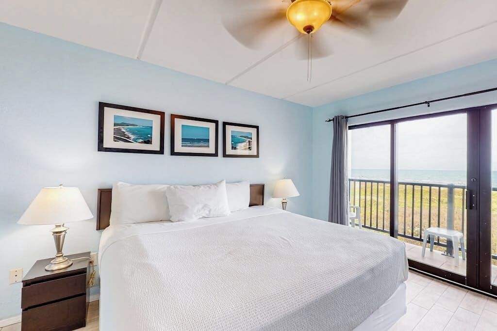 A hotel room with a bed, two bedside lamps, a ceiling fan, and a balcony with a view of the ocean.