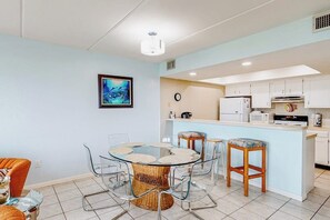 A modern kitchen with white cabinets, a central island with stools, and a dining area with a glass table and chairs. - Continental #201 - Gulf-Front with Beach Access (South Padre Island)