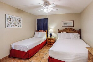 A hotel room with two single beds, a ceiling fan, a framed botanical print, a window with a blue curtain, and a small bedside table with a lamp. - Suntide II Condominiums #402 (South Padre Island)