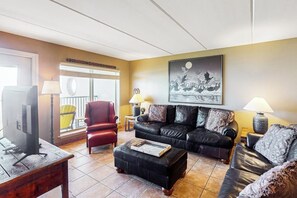 A living room with a sofa, ottoman, and a television on a wooden table. - Suntide II Condominiums #402 (South Padre Island)