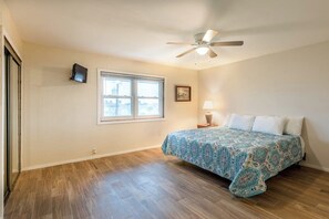 Un dormitorio con cama, ventilador de techo, ventana, televisor montado en la pared y lámpara de noche.