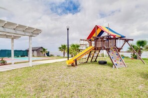 A playground with a slide and climbing net, surrounded by grass and palm trees. - Las Joyas Bayview Home #202 (Port Isabel)