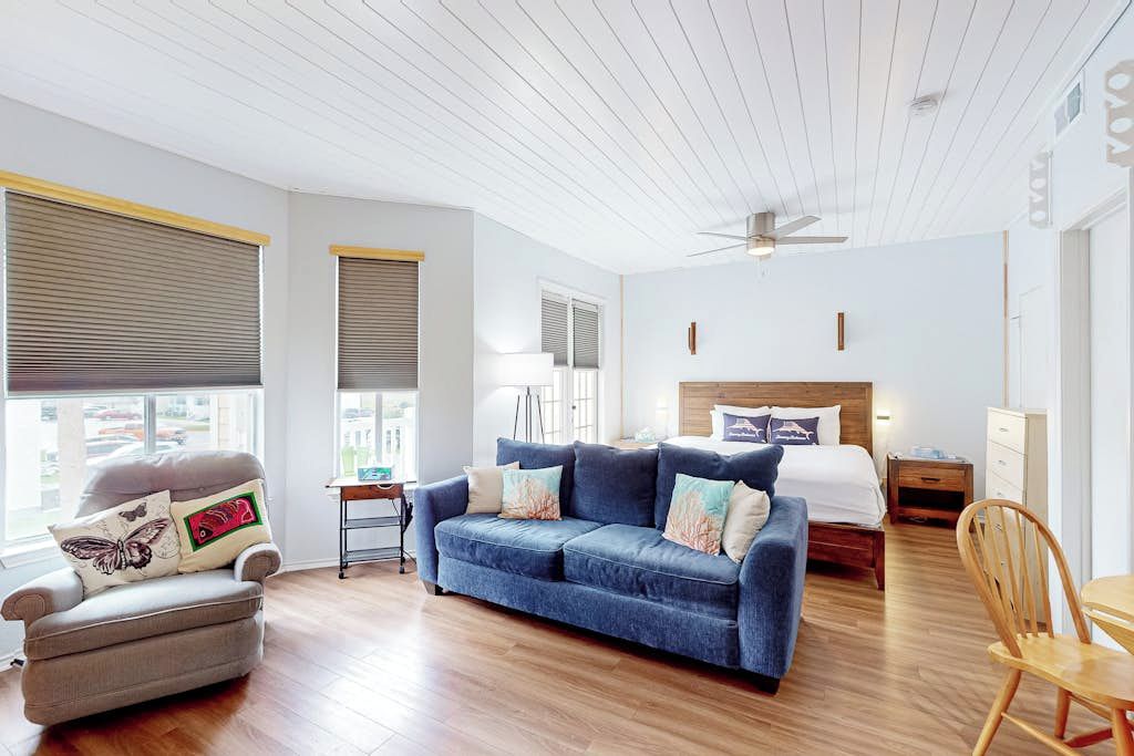 A modern room with a blue sofa, a grey armchair, a wooden dining table, and a bed with a wooden headboard.