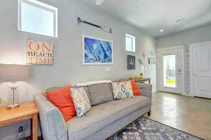 A living room with a grey sofa, a wooden side table, and a patterned rug. There are two windows and a door in the background. - Beachy Keen - Dog-Friendly with Shared Pool (Port Aransas)