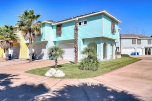 A two-story house with a garage, a front lawn with palm trees, and a clear blue sky. - Beachy Keen - Dog-Friendly with Shared Pool (Port Aransas)