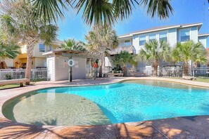 A swimming pool with a curved edge, surrounded by palm trees and a building with a balcony. - Beachy Keen - Dog-Friendly with Shared Pool (Port Aransas)