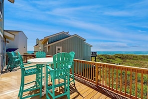 A wooden deck with outdoor furniture, a view of the ocean, and a building with a blue exterior. - Lost on the Sea | Lost Colony 3 - Gulf Views, Pool, Walk to Beach (Port Aransas)