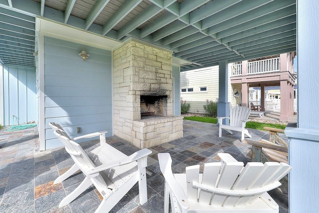 An outdoor patio with a stone fireplace, white Adirondack chairs, and a covered area with a metal roof.
