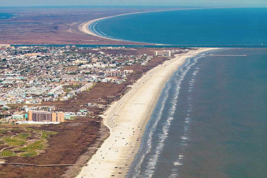 An aerial view of a coastal city with a long beach, dense residential area, and a prominent building complex.