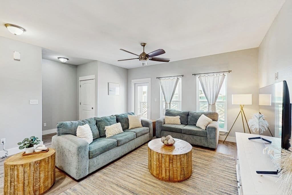 A modern living room with two grey sofas, a ceiling fan, and a large window with curtains.