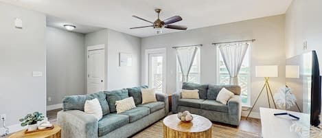 A modern living room with two grey sofas, a ceiling fan, and a large window with curtains.
