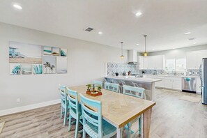 A modern kitchen with white cabinets, a central island, and a dining area with a wooden table and blue chairs. - Shoreline Retreat - Pool & Hot Tub, Walk to Beach (Port Aransas)