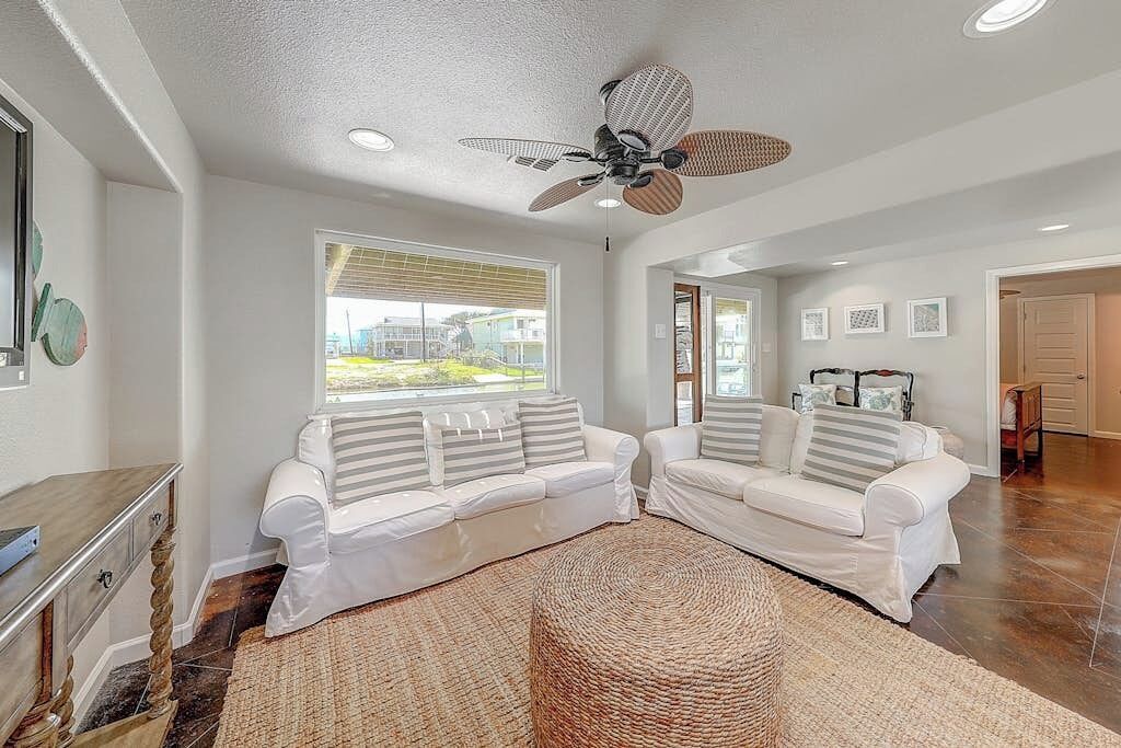 A living room with two white sofas, a woven rug, a ceiling fan, and a view of the outside.