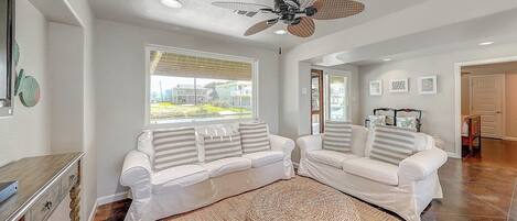A living room with two white sofas, a woven rug, a ceiling fan, and a view of the outside.