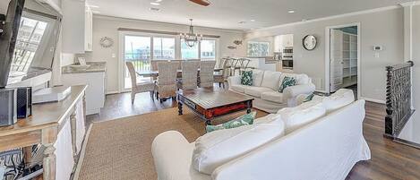 A spacious living room with a white sofa, a dining area with wicker chairs, a ceiling fan, and a view through the glass door.