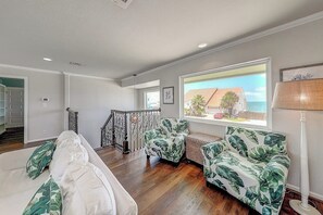 Ein großzügiger Wohnbereich mit einem weißen Sofa, zwei gemusterten Sesseln, einem großen Fenster mit Ausblick und einer Treppe, die in eine obere Ebene führt.