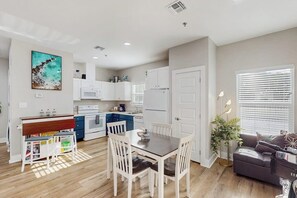 A modern kitchen with white cabinets, a dining area with a wooden table and chairs, and a living room with a sofa and a plant. - Three Palms By The Bay (Rockport)