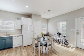 A modern kitchen with white cabinets, a dining table, chairs, and a bicycle. - Three Palms By The Bay (Rockport)