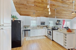 A modern kitchen with white cabinets, a black refrigerator, stainless steel appliances, and wooden ceiling beams. - Bay Breeze House - Waterfront, Dock & Hot Tub (Rockport)