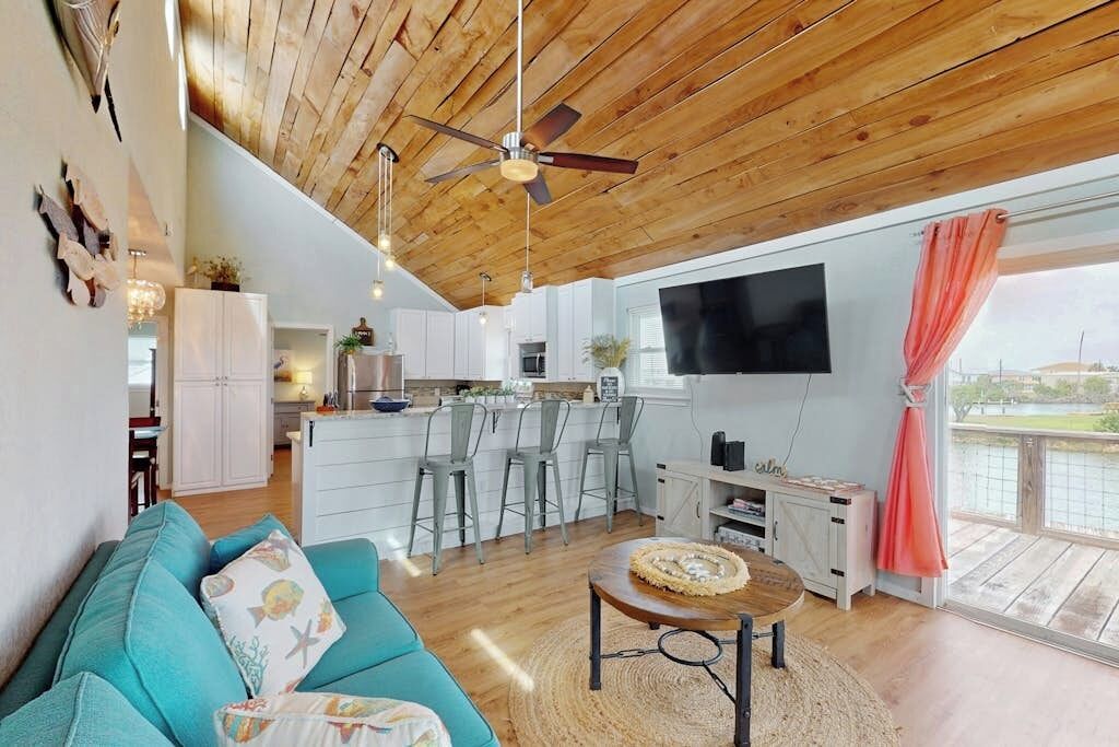 A modern living space with a wooden ceiling, a kitchen area, a seating area with a sofa, and a large window with a view.