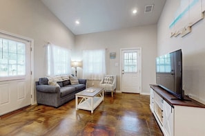 A living room with a sofa, coffee table, TV, and a door. - Casa Azul - Fenced Yard, Fire Pit & Dogs OK (Rockport)
