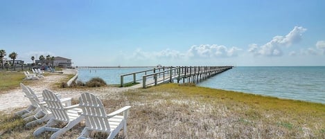 Une plage avec une jetée en bois qui s’avance dans l’eau, une rangée de chaises de plage blanches et un bâtiment en arrière-plan.