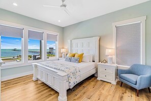 A bedroom with a large bed, a nightstand, a chair, and a view of the ocean through large windows. - Water Street Pearl - Gulf Views & Outdoor Kitchen (Rockport)
