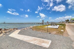 A coastal walkway with a concrete path leading to a grassy area with a bench and a signpost. - Water Street Pearl - Gulf Views & Outdoor Kitchen (Rockport)