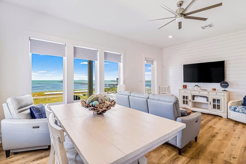 A modern living room with a large window, a ceiling fan, a flat-screen TV, and a dining table with a decorative bowl.