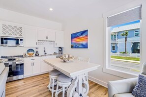 A modern kitchen with white cabinetry, a dining table with four chairs, and a large window with a view of a blue house and palm tree. - Water Street Pearl - Gulf Views & Outdoor Kitchen (Rockport)