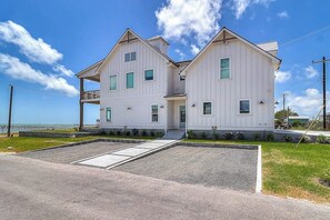 A two-story white building with a balcony on the upper floor, a driveway, and a grassy area in front. - Water Street Pearl - Gulf Views & Outdoor Kitchen (Rockport)