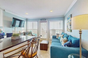 A modern living room with a blue sofa, a dining area with a bar, and a TV mounted on the wall. - Seascape Condos - Day Break at the Seascape #2219 (Galveston)